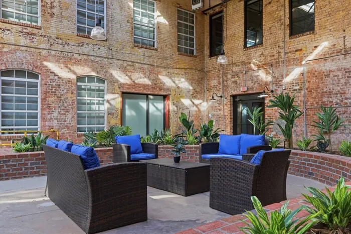 Charming industrial-style courtyard featuring cozy wicker seating with vibrant blue cushions, surrounded by lush greenery and rustic brick walls.