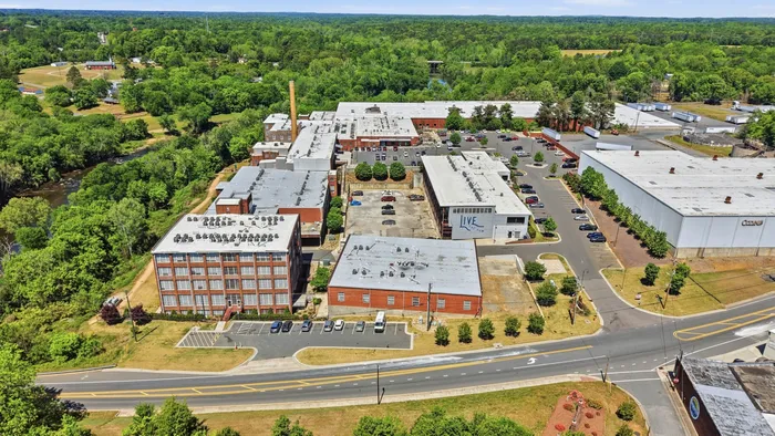 Aerial view showcasing a vibrant residential and commercial area surrounded by lush greenery and a meandering river, offering ample parking and modern architectural design.