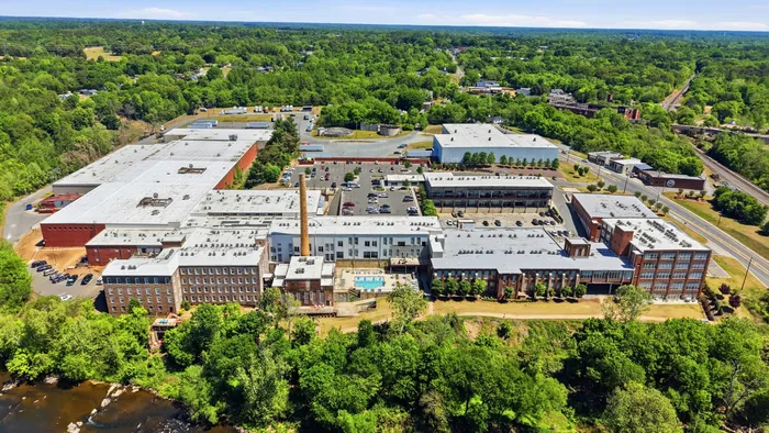 Expansive aerial view showcasing a vibrant complex with industrial-style buildings surrounded by lush greenery and a sparkling pool, offering modern living amidst nature.