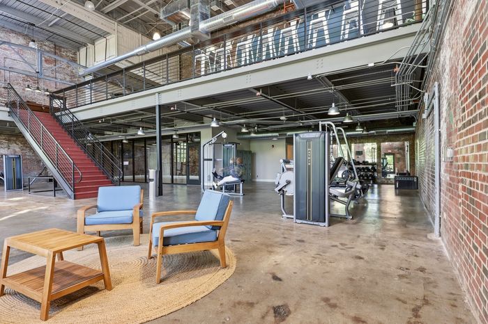 Modern industrial-style gym with exposed brick walls, high ceilings, and a blend of fitness equipment and comfortable seating area.