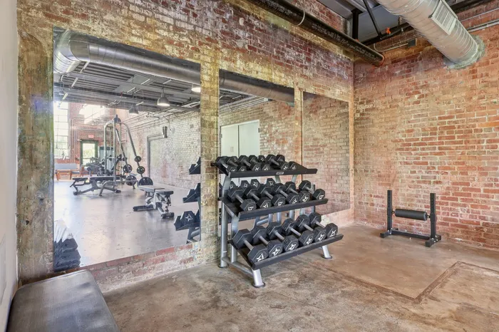 Industrial-style gym with exposed brick walls and polished concrete floors, featuring a full set of dumbbells and modern equipment.