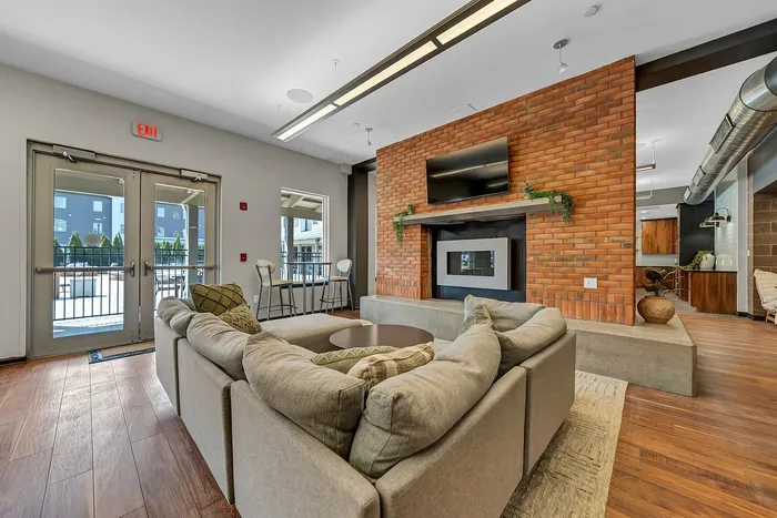 Chic community apartment living room featuring a cozy sectional sofa, a brick feature wall with a modern gas fireplace and TV, and an inviting open layout with abundant natural light. View of the patio and hot tub.