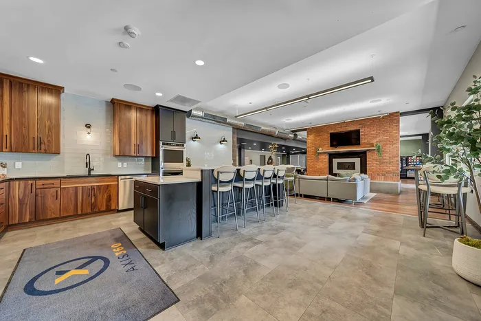 Modern open-concept kitchen and living area featuring sleek wooden cabinets, island seating, and a cozy brick gas fire place. Luxury apartment complex event space.