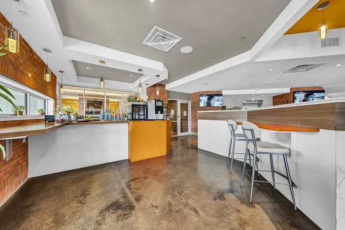 Modern cafe in luxury apartment lobby with sleek design, featuring stylish pendant lighting, a vibrant orange counter, and cozy bar seating against a backdrop of exposed brick walls. Free coffee bar and waffle bar supplies.