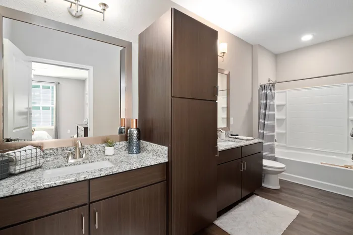 Chesterfield, MO Apartments Near Chesterfield Mall - Bathroom With Granite Countertops, Wood-Style Flooring, Two Separated Vanities, Dark Wood-Style Cabinets, And A Shower With Tub And Small Built-In Shelves.