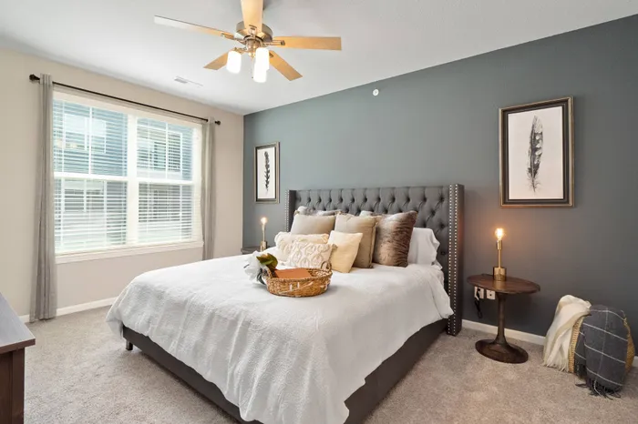 Apartments in Chesterfield, MO - The Parq at Chesterfield - Spacious Bedroom with Carpet Flooring, Large Bed, Cushioned Bed Headboard, Nightstands with Lamps, Ceiling Fan, Wall Art, and Large Window with Blinds.
