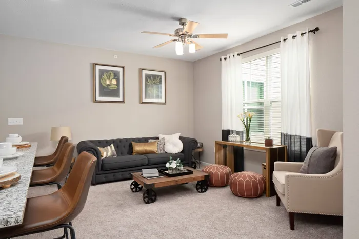 Pet-Friendly Apartments in Chesterfield, MO - The Parq at Chesterfield - Living Room with Carpet Flooring, Couch, Ottoman Stools, Armchair, Coffee Table, Side Tables, Ceiling Fan, Window with Blinds, and View of Breakfast