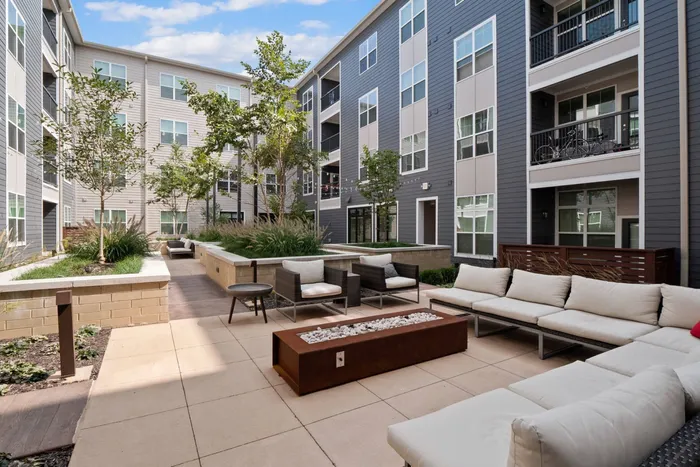 2-Bedroom Apartments in Chesterfield, MO - The Parq at Chesterfield - Courtyard with Plush Seating, Firepit, String Lighting, Maintained Landscaping, and Apartment Buildings in the Background.