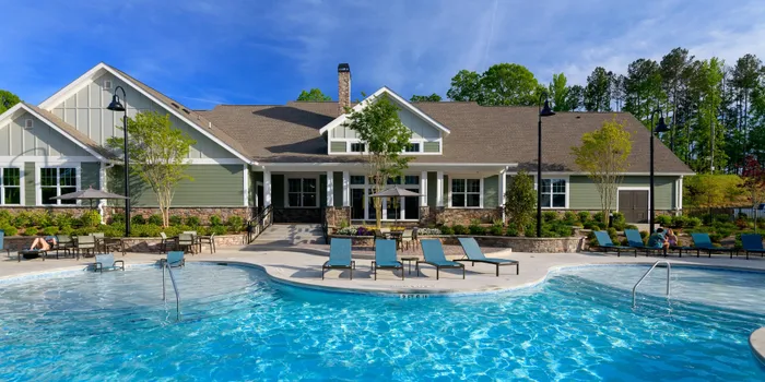 Apartments in West Athens, GA - Lullwater at Jennings Mill - Resort Style Pool With Lounge Chairs, Sundeck Seating, Landscaped Clubhouse Exterior, And Bright Blue Sky.