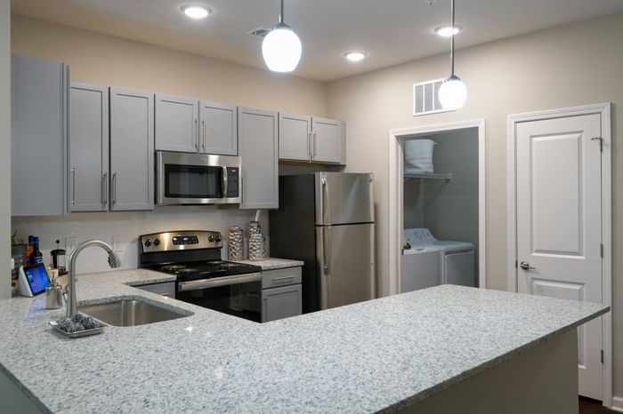 1 Bedroom Apartments in Athens, GA - Lullwater At Jennings - Kitchen With Granite Countertops, Subway Tile Backsplash, Gray Shaker Cabinetry With Brushed Nickel Pulls, Stainless Steel Appliances, Pendant Light Fixtures, And View Of Laundry Closet With Washer And Dryer.