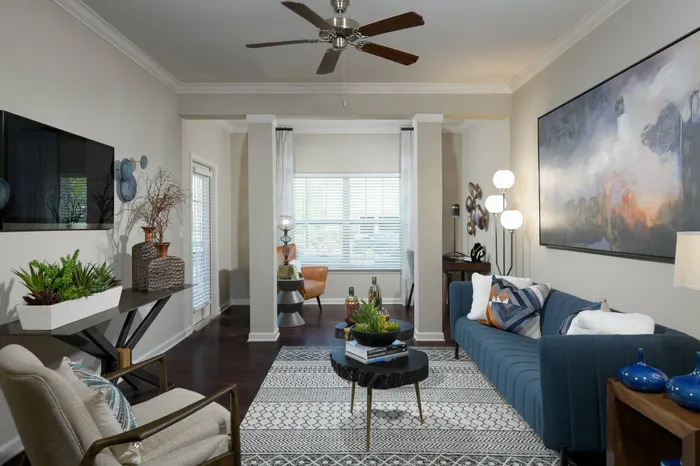 West Athens, GA, Apartments - Lullwater at Jennings Mill - Living Room With Blue Sofa, Wall-Mounted Television, Ceiling Fan, Large Windows, Patterned Area Rug, And Modern Accent Decor.
