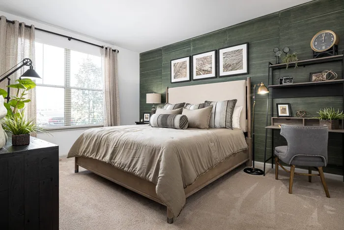 Pet-Friendly Apartments in Buford, GA - The Harrison at Braselton - Bedroom with Carpet Flooring, Large Bed, Desk, Shelves, Nightstand, Lamps, Armchair, Dresser, Clock, Wall Art, and Large Windows.