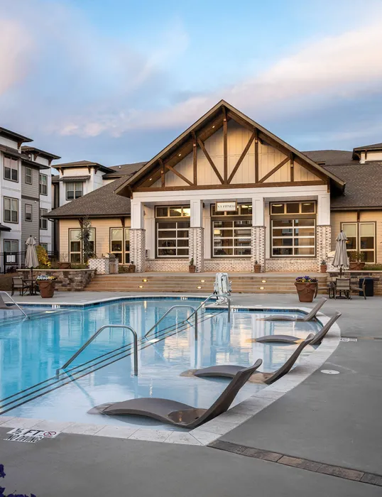 1-Bedroom Apartments in Buford, GA - The Harrison at Braselton - Swimming Pool with In-Water Loungers, Seating with Closed Umbrellas, and View of the Clubhouse and the Apartments.