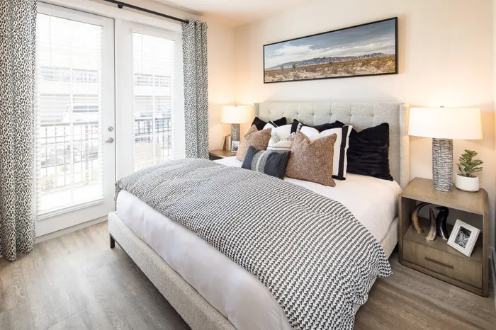 Gainesville, GA, Apartments - The Mill at New Holland - Bedroom with Wood-Style Plank Flooring, Large Bed, Nightstands with Lamps, Wall Art, and Double Glass Doors with Blinds and Access to Patio.