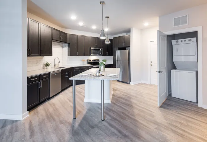 Apartments in Gainesville, GA for Rent - The Mill at New Holland - Kitchen with Wood-Style Plank Flooring, Kitchen Island with Pendant Lights, Granite Countertops, Designer Ceramic Tile Backsplash, Stainless-Steel Appliances, Wood-Tone Cabinets, and View of Laundry Closet with Washer and Dryer.