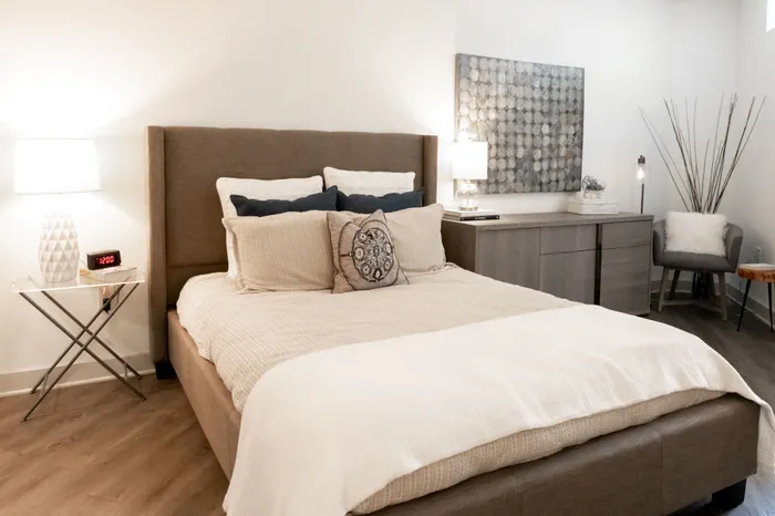 One BR Apartments in Downtown Birmingham AL - Park 35 on Clairmont - Bedroom with Wood Flooring, a Bed, Wooden Dresser, Nightstand with Lamp, Corner Chair, Mounted Wall Art, and White Walls