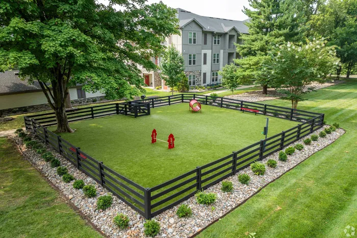 Lush dog park with agility equipment surrounded by apartment buildings and trees.