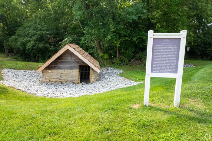 A quaint stone springhouse surrounded by lush greenery and a serene landscape with a nearby informational sign.