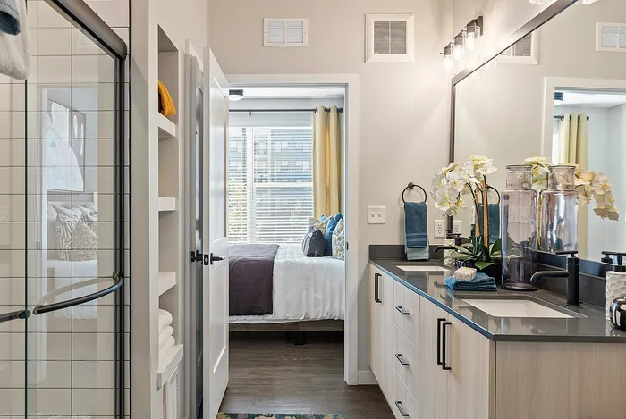 Contemporary apartment bathroom with dual sinks, elegant decor, and a view into a brightly lit bedroom featuring large windows.