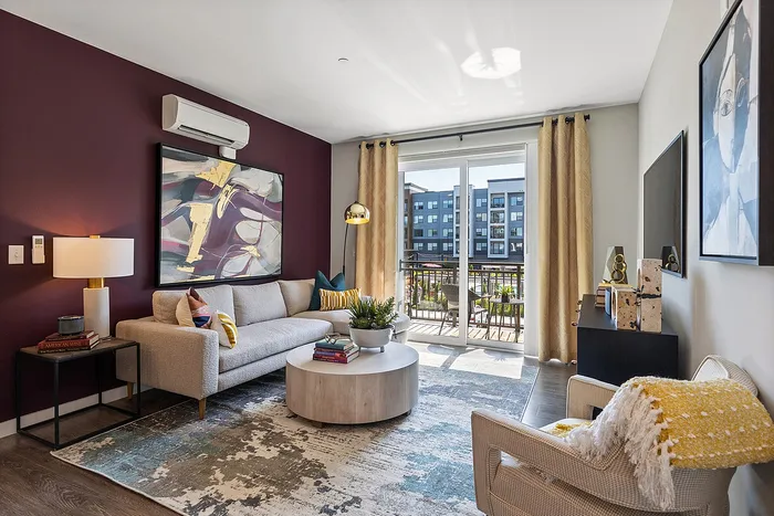 Bright and stylish living room featuring a cozy beige sofa, elegant artwork on a striking maroon accent wall, and access to a sunny balcony with modern sliding glass doors.