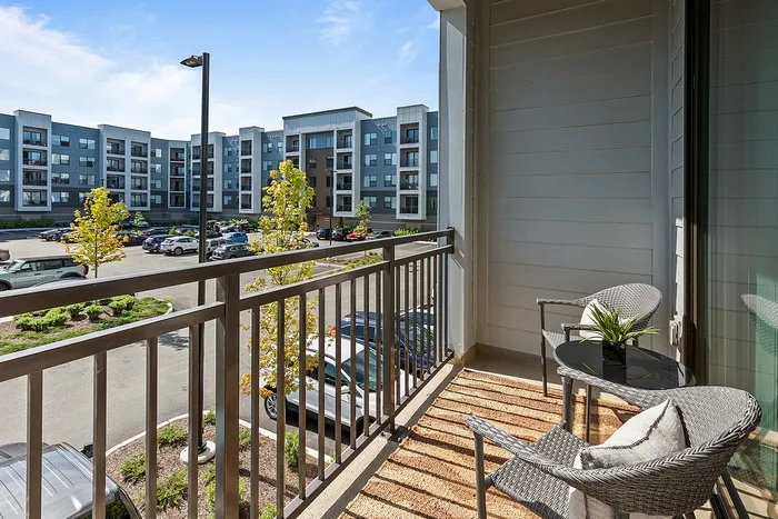 Inviting apartment balcony with stylish wicker furniture and a lush potted plant, offering a picturesque view of modern residential buildings and a vibrant courtyard.