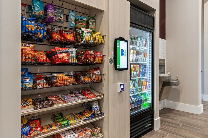 Modern snack and beverage station featuring a variety of chips, candies, and drinks in a well-lit, sleek display with a digital payment kiosk.