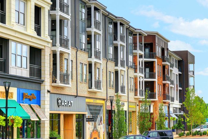Vibrant urban apartment complex featuring modern architecture with retail stores on the ground floor, lush greenery, and ample natural light under a bright blue sky.