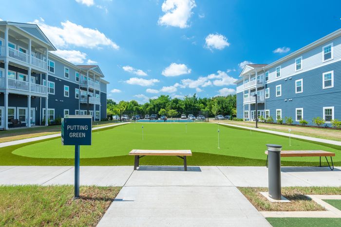 Modern apartment complex with vibrant blue exterior featuring spacious balconies, surrounded by lush greenery and a well-maintained putting green under a clear blue sky.