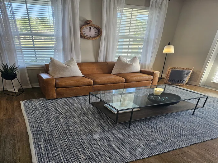 Inviting living room with elegant brown leather sofa, stylish glass coffee table, decorative rug, and natural light from large windows.