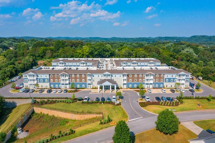Aerial view of a modern apartment complex surrounded by lush green trees, featuring stylish three-story buildings with ample parking and scenic landscape.