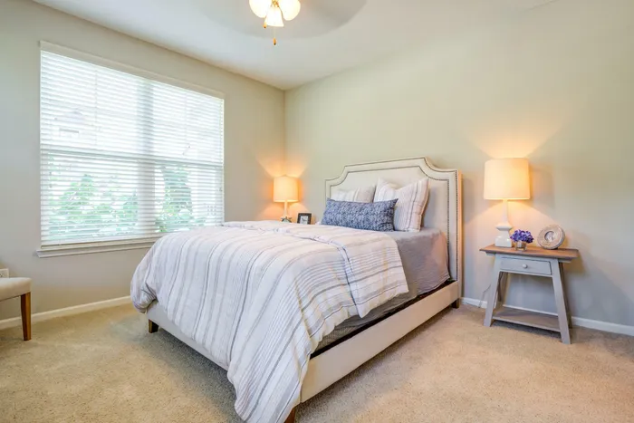 Bright and cozy bedroom featuring a plush bed with elegant striped bedding, soft carpet, and warm ambient lighting.
