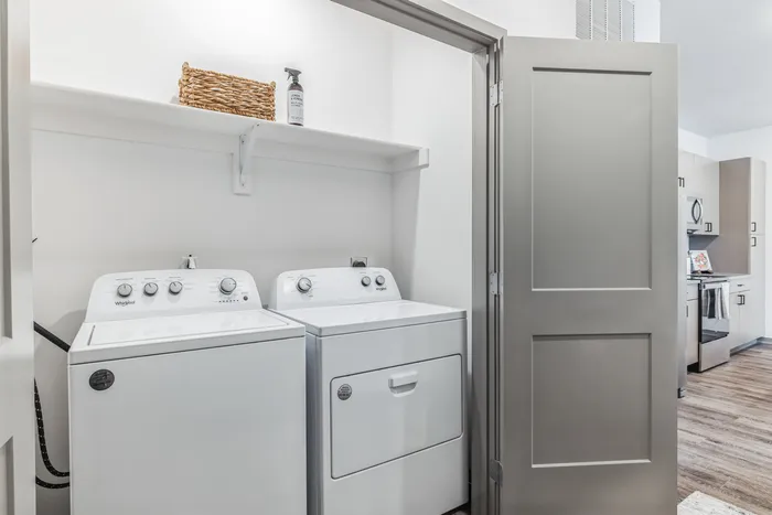 Modern apartment laundry room with sleek washer and dryer, adjacent to stylish kitchen with wood flooring.