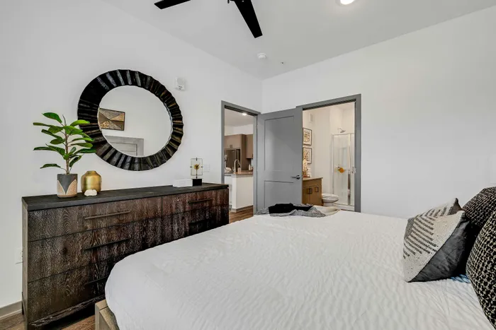 Chic bedroom with modern decor, featuring a stylish circular wall mirror, elegant dark wood dresser, and a glimpse into a contemporary bathroom.