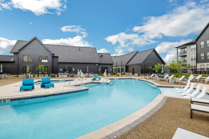 Luxurious apartment complex featuring a stunning outdoor pool area with modern lounge chairs and vibrant blue seating, all under a clear blue sky.