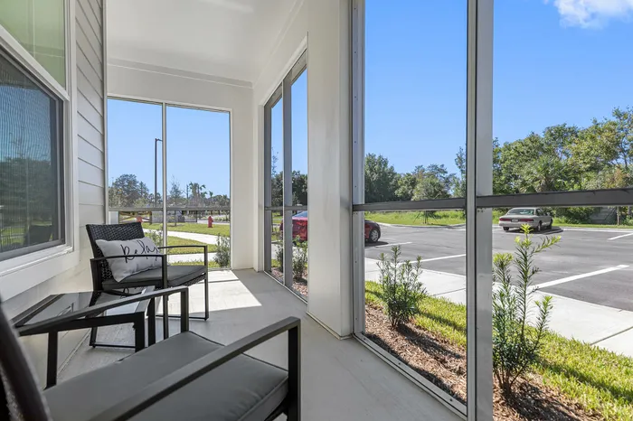 Charming screened-in porch with cozy seating, offering tranquil views of lush greenery and a sunny parking area.