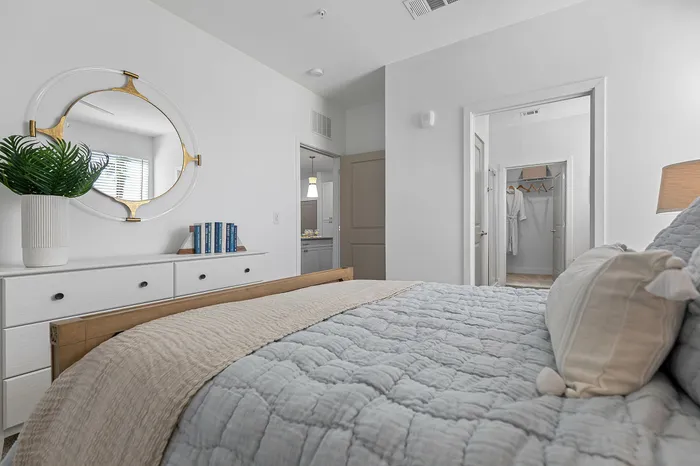 Bright and inviting bedroom with modern decor features a comfortable bed, stylish dresser with books, and a contemporary mirror; open door leads to a walk-in closet and ensuite bathroom.