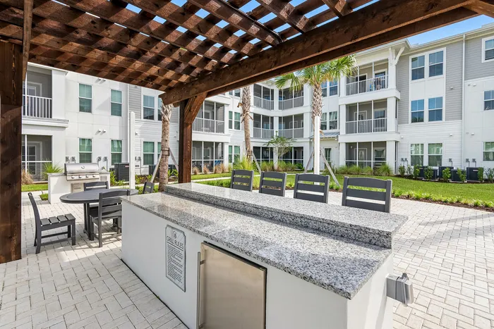 Chic outdoor lounge area featuring a modern BBQ setup under a rustic pergola, surrounded by lush landscaping and stylish apartments.