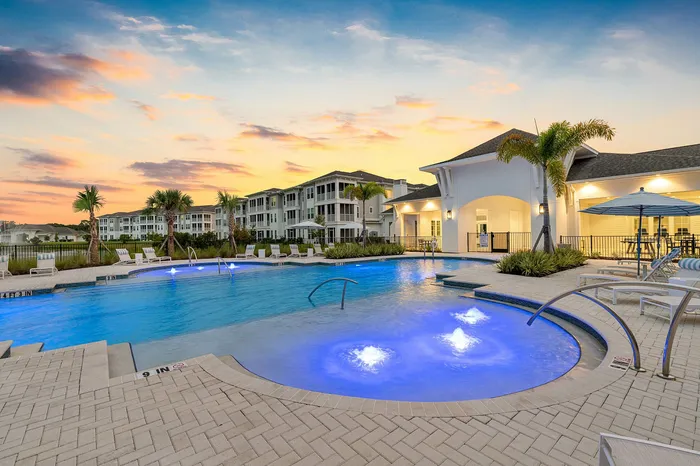 Luxurious apartment complex featuring a stunning illuminated pool area with elegant lounge chairs, set against a beautiful sunset.