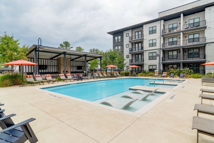 Modern apartment complex with a luxurious outdoor swimming pool, surrounded by stylish lounge chairs and vibrant orange umbrellas, ideal for relaxation and leisure.