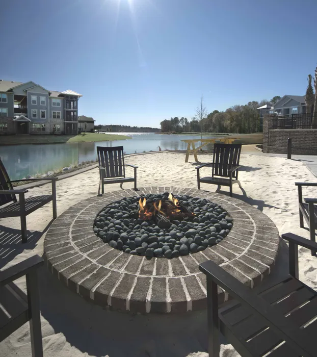 Inviting outdoor fire pit area with elegant brick design, surrounded by comfortable seating, overlooking a serene lake on a sunny day.