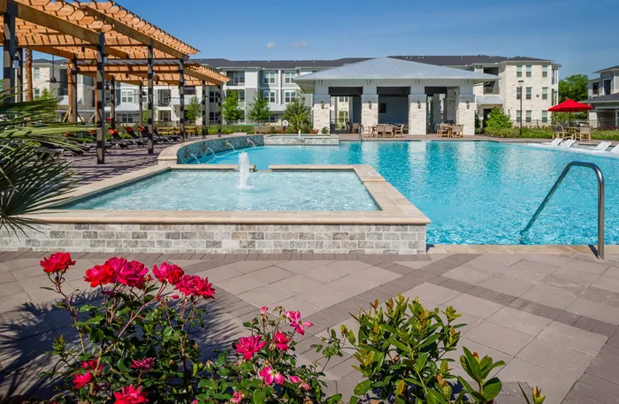 Luxurious apartment complex with a sparkling outdoor pool, stylish pergola, and vibrant landscaping featuring blooming red flowers.