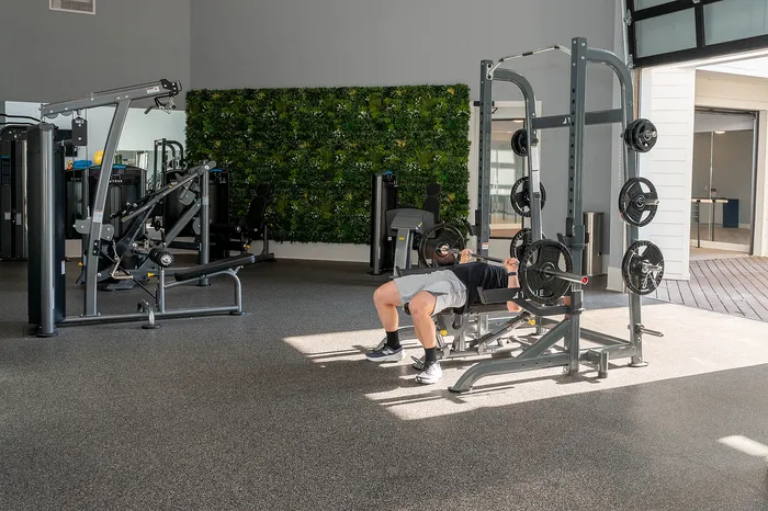 Modern fitness center with natural light and a vibrant green wall, featuring advanced weightlifting equipment and a person bench pressing.