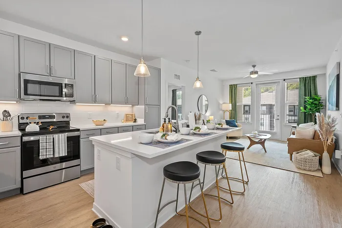 Modern open-concept kitchen and living area with sleek gray cabinets, stylish bar stools, and bright natural light through large windows.