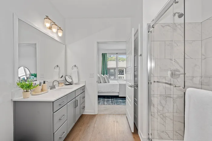 Sleek modern bathroom with elegant double vanity, large mirror, and shower, opening to a bright bedroom with stylish decor.