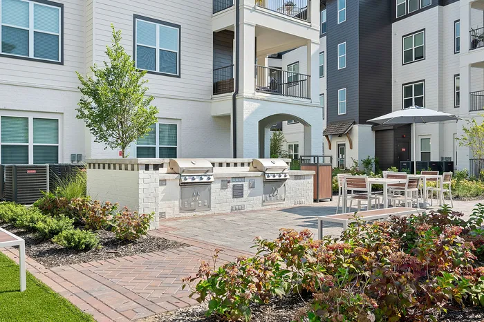 Charming apartment complex featuring a modern outdoor BBQ area, elegant seating with umbrellas, and lush landscaping.