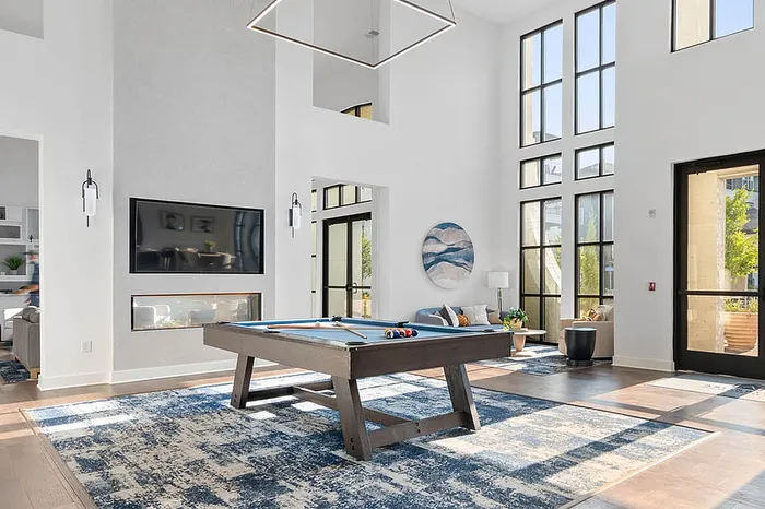Spacious and modern recreation room with high ceilings, featuring a pool table, fireplace, and large windows for abundant natural light.