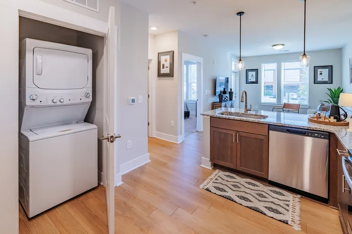 Modern apartment with a sleek kitchen featuring a granite island, stainless steel appliances, and a built-in washer and dryer, leading to a bright living area.