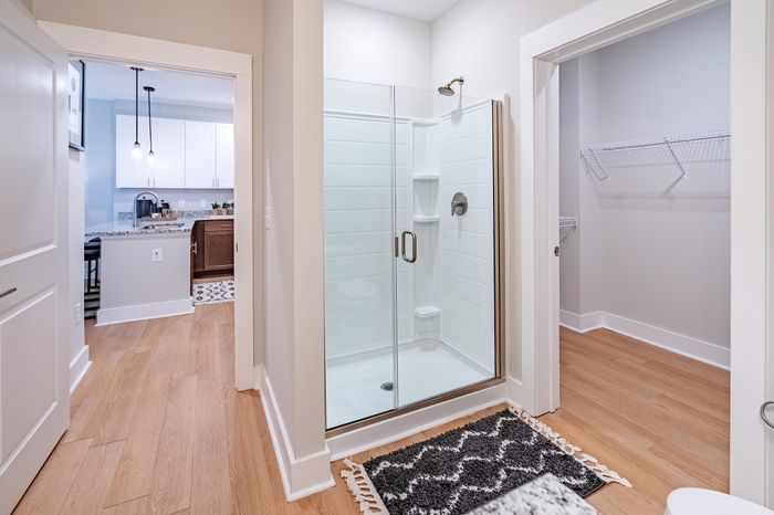 Modern apartment bathroom with a glass-enclosed shower, hardwood floors, and a glimpse of the stylish kitchen in the background.