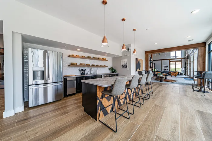 Modern kitchen and living area featuring a sleek kitchen island with geometric design and contemporary pendant lighting. Spacious open floor plan with stylish bar stools and ample natural light.