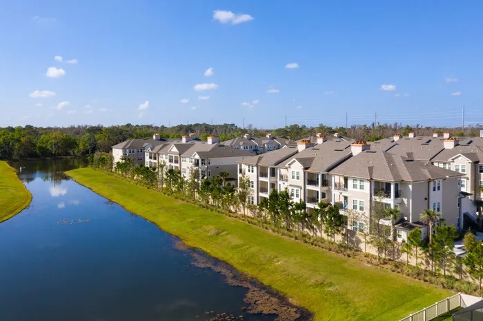 Charming apartment complex nestled by a serene lake, featuring modern architecture and lush greenery under a sunny blue sky.